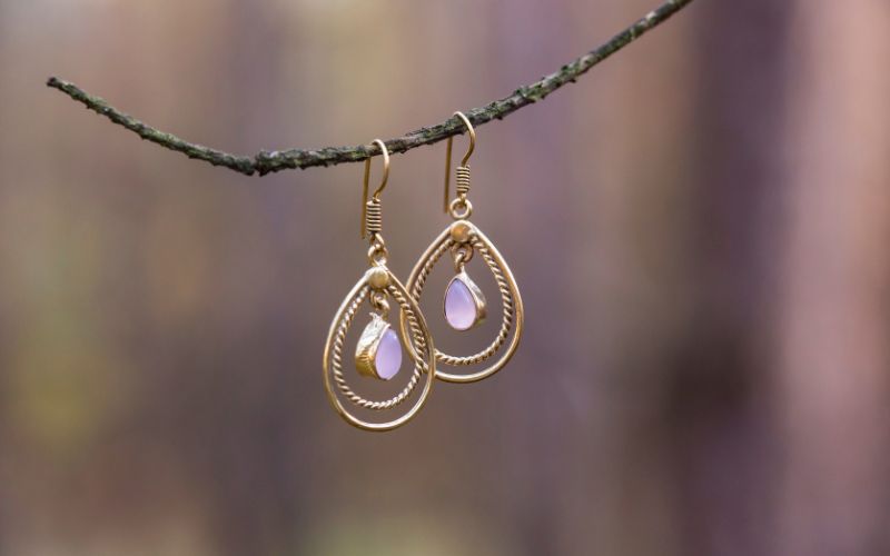 Boucle d'oreille goutte d'eau détournée, couleur doré et blanc flou, accrochées a une fine branche d'arbre, l'arrière plan est flou