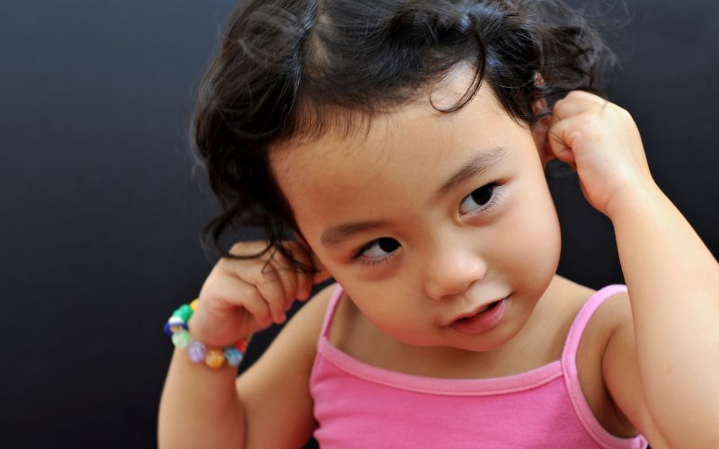 Bébé qui porte un débardeur rode, qui porte un bracelet en perle plasqtique, qui se touche les oreilles