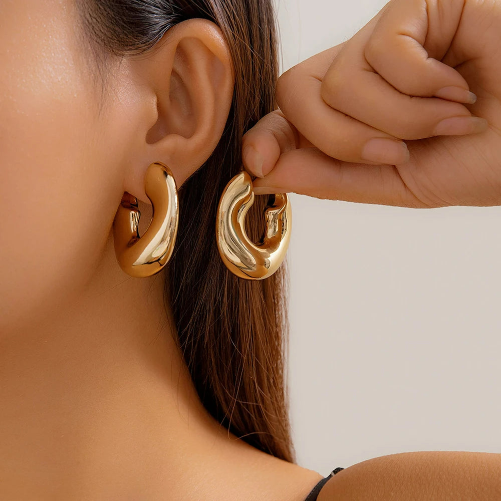 Boucle D'oreille Créole Épaisse Originale Dorée portée par une femme, l'autre boucle est entre ses doigts, elle la présente à côté de celle à son oreille.