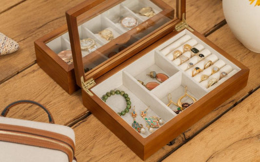 Belle boîte à bijoux en bois avec vitre en verre, posée devant un miroir sur un meuble en bois. Pleins de bijoux sont bien rangés à l'intérieur.