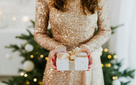 Femme en robe de soirée beige brillante a manche longue, tenant un cadeau dans ses mains tendues vers nous et un sapin en arrière plan flou.
