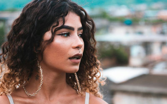 Femme de trois quart, portant de très grande créoles dorées, avec les cheveux détaché et ondulés. Le fond derrière elle est flou 