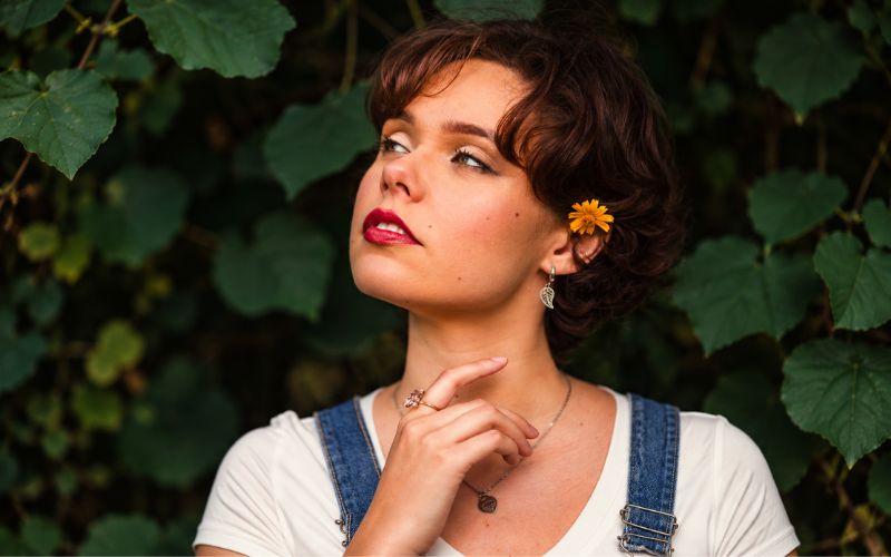 Femme qui regarde légèrement en l'air de trois quart profil, elle porte une boucle d'oreille anneau avec pendentif feuille. Derrière elle des feuille verte de lierre  Image