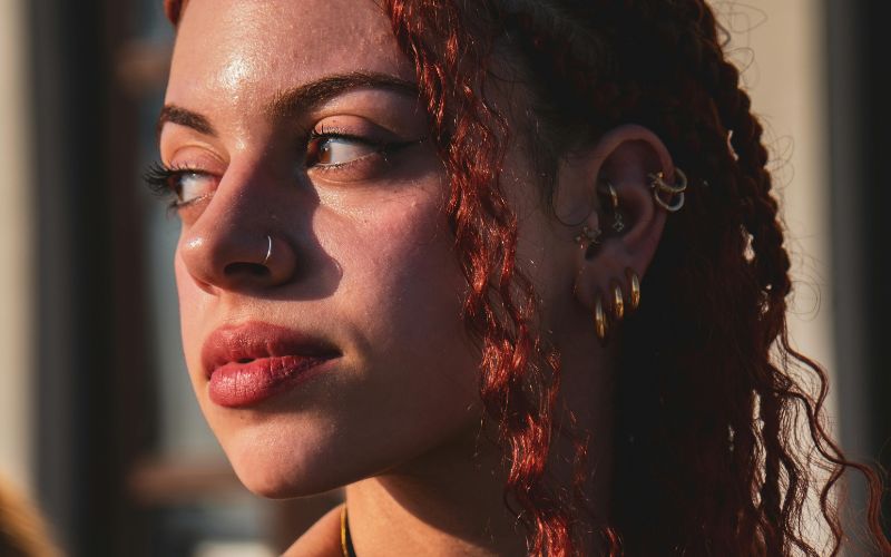 Femme de trois quart, on ne voit que sa tête et ses cheveux, elle a un piercing au nez et un lot de 3 boucles d'oreille créoles dorées et des boucles d'oreille au cartilage. Image