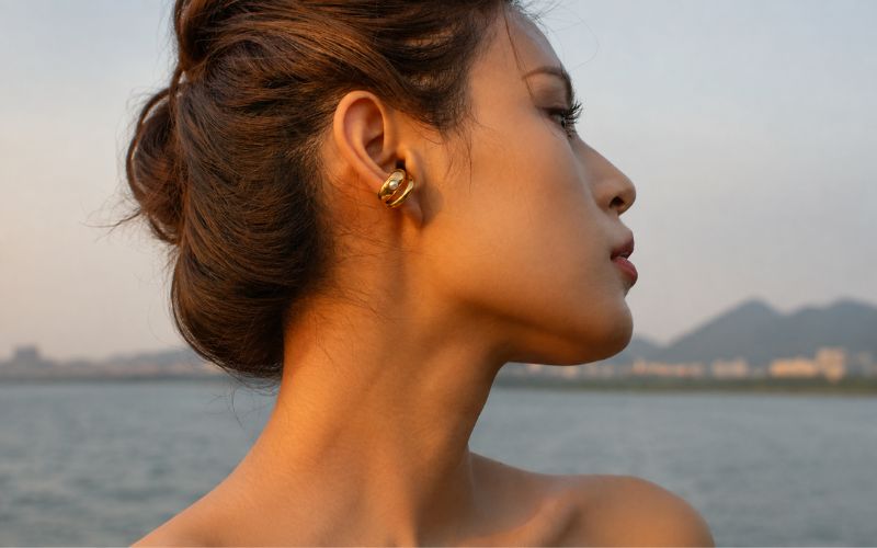 Femme devant un plan d'eau, la tête de trois-quarts, les cheveux attachés en chignon effet coiffé/décoiffé, portant une ear cuff anneau doré double avec une perle blanche posé sur le plus gros anneau Image