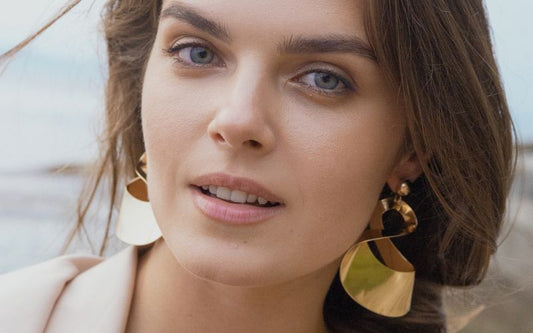 Femme portant des grande boucle d'oreilles pendantes dorées en acier inoxydable. On voit la mer derrière elle.