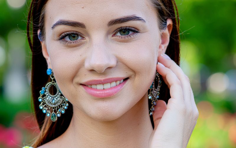 Femme souriante, de face, portant de grandes boucles d'oreille turquoise et argentées, sa main gauche touche son lobe d'oreille et la boucle d'oreille.