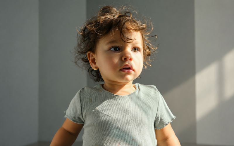 Petite fille, porte un t-shirt gris, marche et on voit des boucles d'oreille clip doré. le fond un mur gris