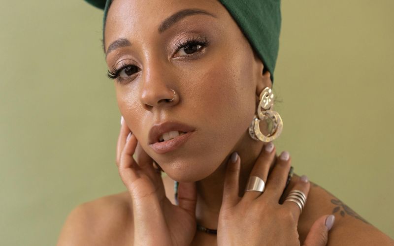 Femme avec maquillage légé, portant une paire de boucle d'oreille clip pendante dorée en forme arrondi, elle porte aussi des bagues et ses mains sont posées sur son coupr et le bas de son visage. le fond derrière elle est vert olive clair.  