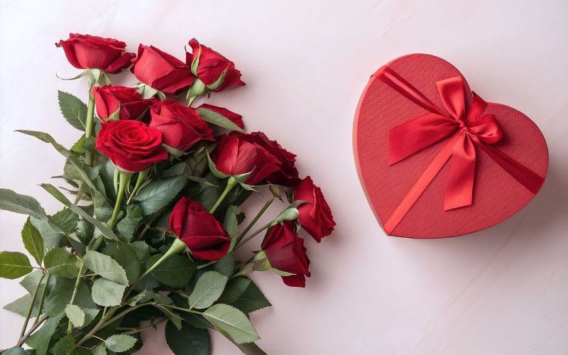 Bouquet de pleins de roses rouges, posées à côté d'une boite cadeau rouge en forme de cœur avec un ruban rouge.