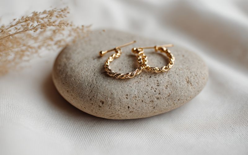 Paire de créole dorée tréssée, présenté sur une jolie caillou gris beige, sur un tissu de Santin beige et un brin de plante séché