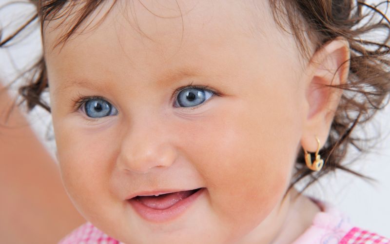 Bébé fille avec de beaux yeux bleus qui sourit, la tête de trois quart, on voit une boucle d'oreille créole à son oreille gauche.