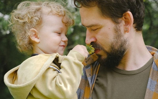 Homme avec son bébé dans les bras qui lui fait sentir une fleur, l'homme porte une boucle d'oreille puce argenté.