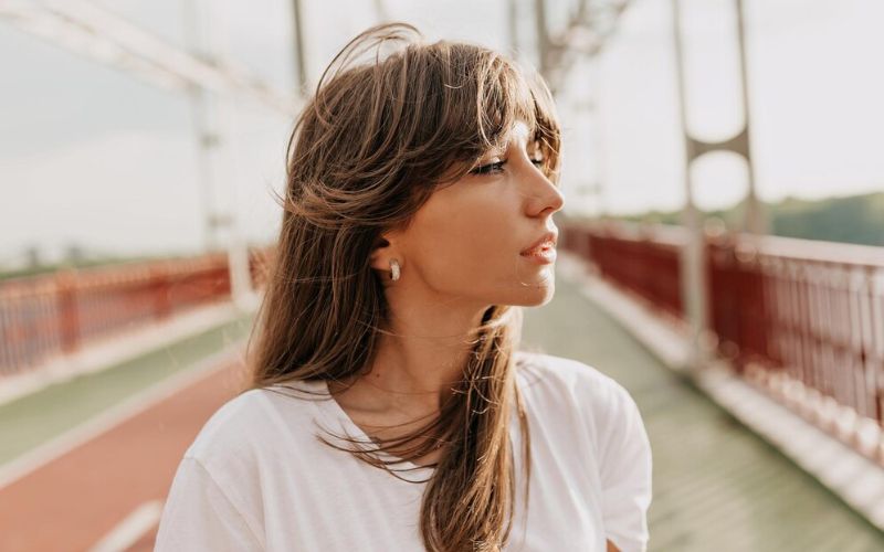 Femme dehors sur un pont, la tête de profil avec les cheveux au vent, on aperçois une jolie boucle d'oreille minimaliste.