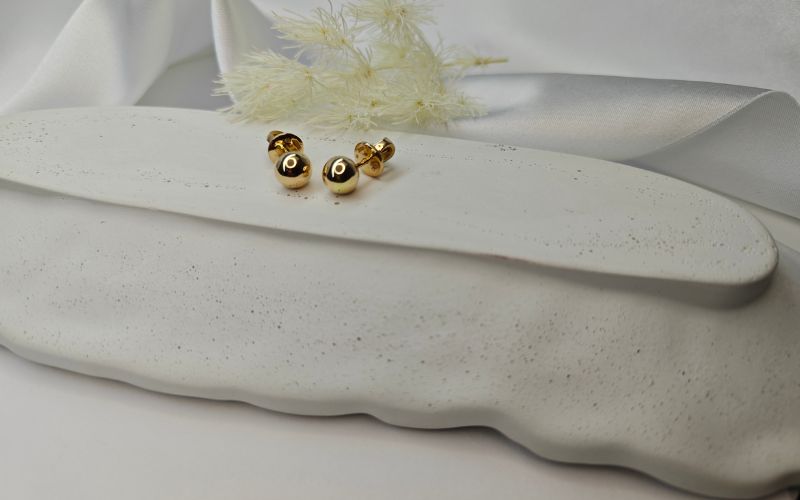 Boucle d'oreille puce perle dorée, mise en valeur sur plat en céramique blanc avec plante séché derrière