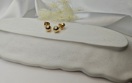 Boucle d'oreille puce perle dorée, mise en valeur sur plat en céramique blanc avec plante séché derrière