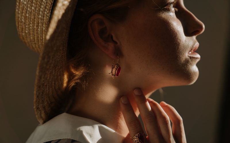 Femme de profil portant un chapeau style paille, face à de la lumière, ses doigts touche son coup près de la boucle d'oreille pendante dorée et rouge qu'elle porte Image