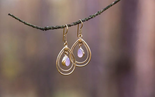 Boucle d'oreille goutte d'eau détournée, couleur doré et blanc flou, accrochées a une fine branche d'arbre, l'arrière plan est flou