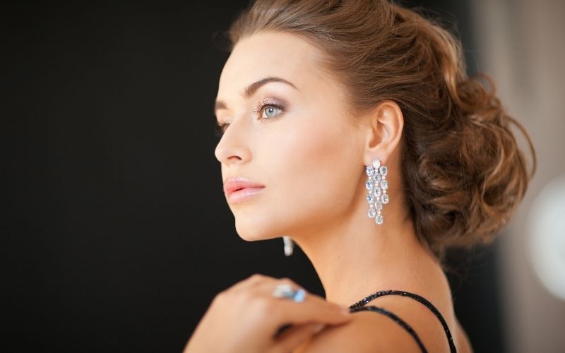 Femme élégant, bien coiffée et maquillé, porte des boucles d'oreilles de luxe pendantes, le fond est flouté.