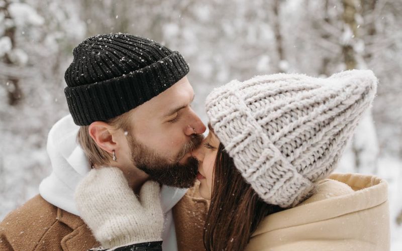 Un homme et une femme, dehors dans la neige, chacun porte un bonnet, l'homme porte une boucle d'oreille anneau et il embrasse le nez de la femme.