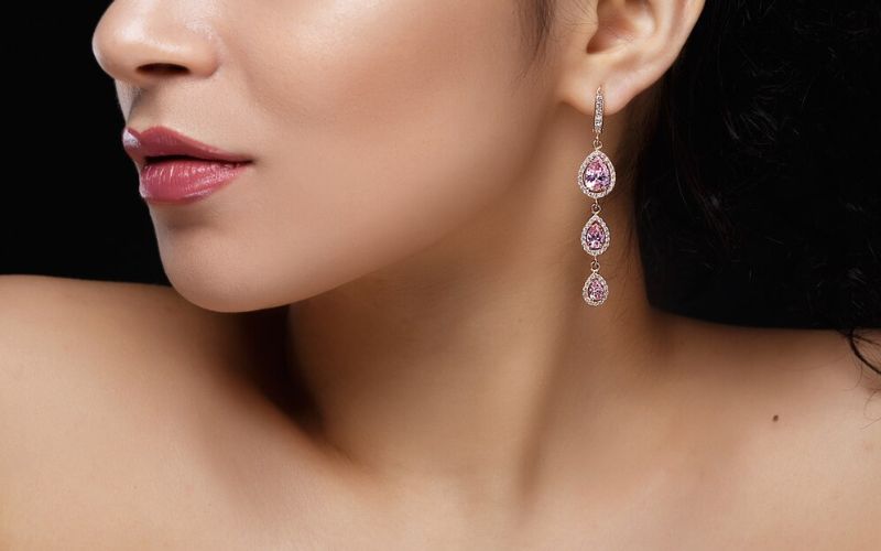Femme portant une boucle d'oreille 3 gouttes d'eau roses et diamant. Elle est de profil, la photo montre le bas du visage et le cou. Image