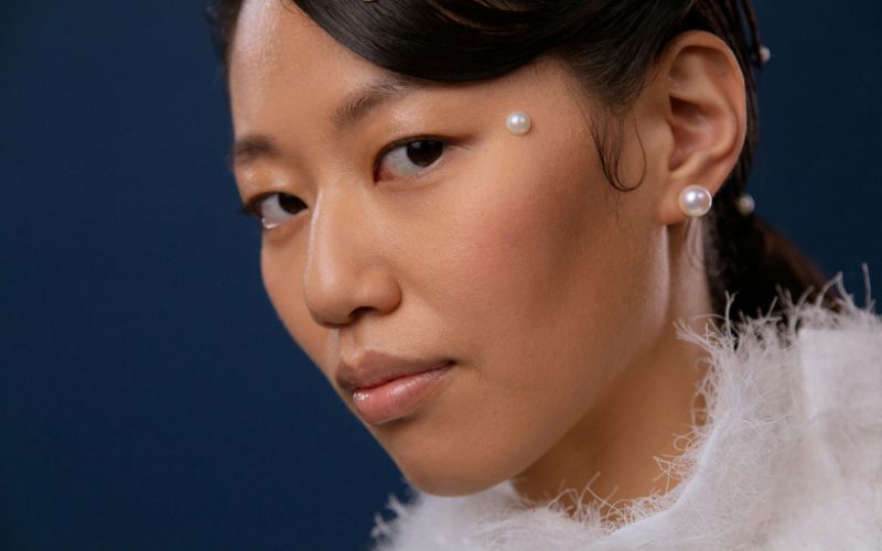 Femme maquillé, coiffée et apprêtée pour un shooting elle porte une boucle d'oreille avec une grosse perle et elle à aussi une perle colée près de l'œil