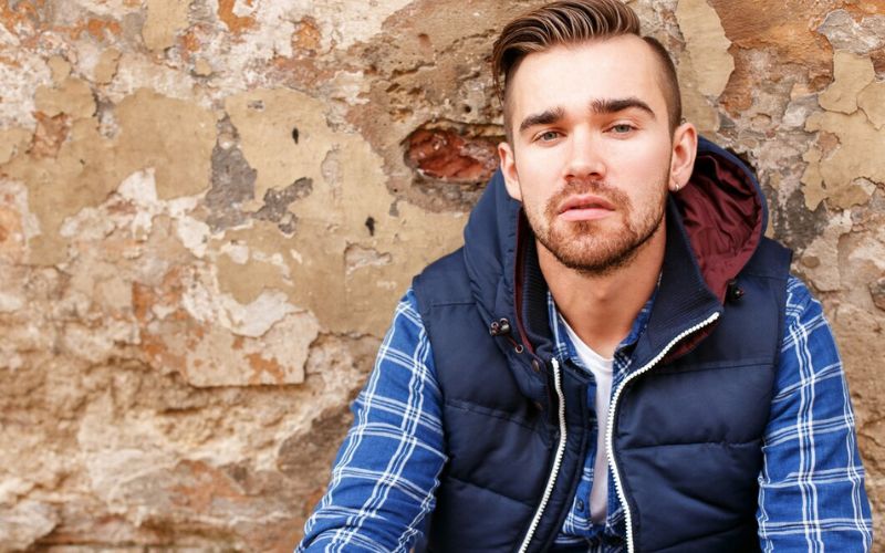 Jeune homme assit contre un mur de pierres, qui porte une chemise bleue à carreaux, une doudoune sans manche et une boucle d'oreille à l'oreille gauche Image