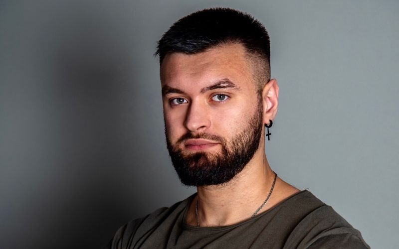 homme avec boucle d'oreille croix sur créole le tout noir, avec un fond gris derrière lui