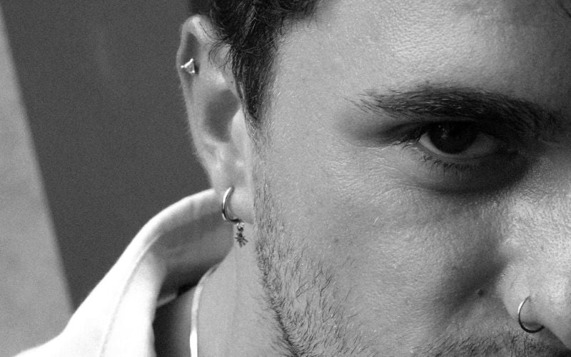 Photo en noir et blanc, d'une homme de près, on ne voit qu'une partie de son visage, il fixe l'objectif, il porte un anneau au nez, une boucle d'oreille puce au cartilage haut de l'oreille et une boucle d'oreille anneau avec pampille au lobe de l'oreille. Image