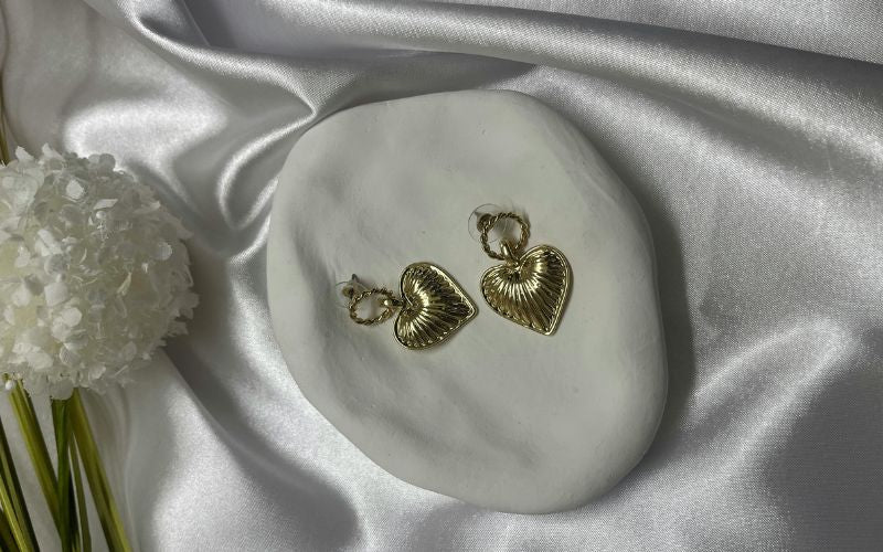 Paire de boucle d'oreille doré pendante cœur, posé sur une présentoir minimaliste pierre blanche, le tout sur un drap en santin blanc et une fleur blanche posé à côté du présentoir. 