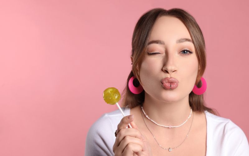 Jeune fille avec un visage rond, qui cligne de l'œil, fais un bisous avec sa bouche, tient une sucette dans sa main, elle porte de grandes créoles rose fuchsia et le fond derrière elle est rose clair.  Image