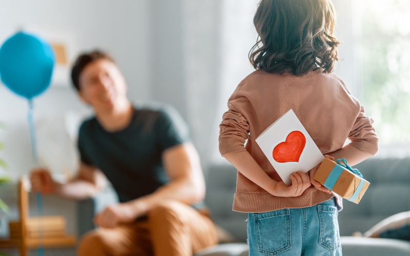Un enfant face à son papa qui cache dans son dos une carte et un cadeau pour la fête des pères.
