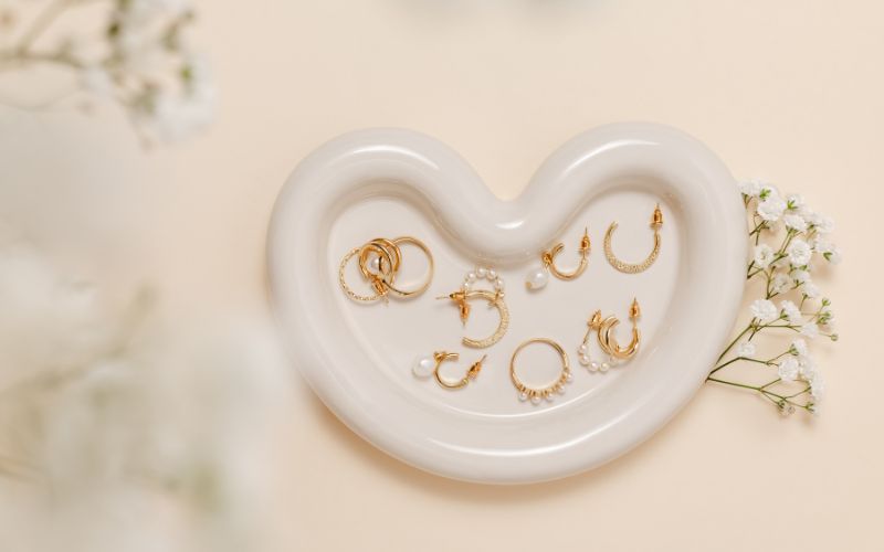 Petit présentoir blanc en forme de cœur sur lequel se trouve pleins de bijoux de piercing pour oreilles et visage. il y à des petites fleur blanche autour.