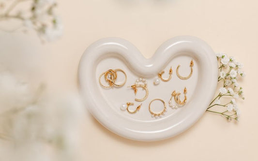 Petit présentoir blanc en forme de cœur sur lequel se trouve pleins de bijoux de piercing pour oreilles et visage. il y à des petites fleur blanche autour.