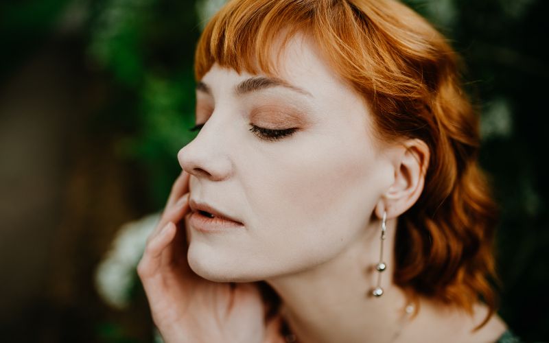 Femme rousse cheveux au carré avec une petite frange, qui se tien le visage dans une de ses main de trois quart, on voit une boucle d'oreille pendante avec des perle en argent