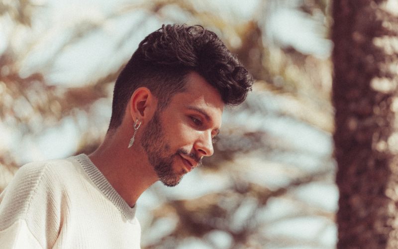 Homme brun les cheveux plutôt court, dehors avec des arbres derrière lui, il porte deux boucle d'oreilles, sur l'oreille droite Image