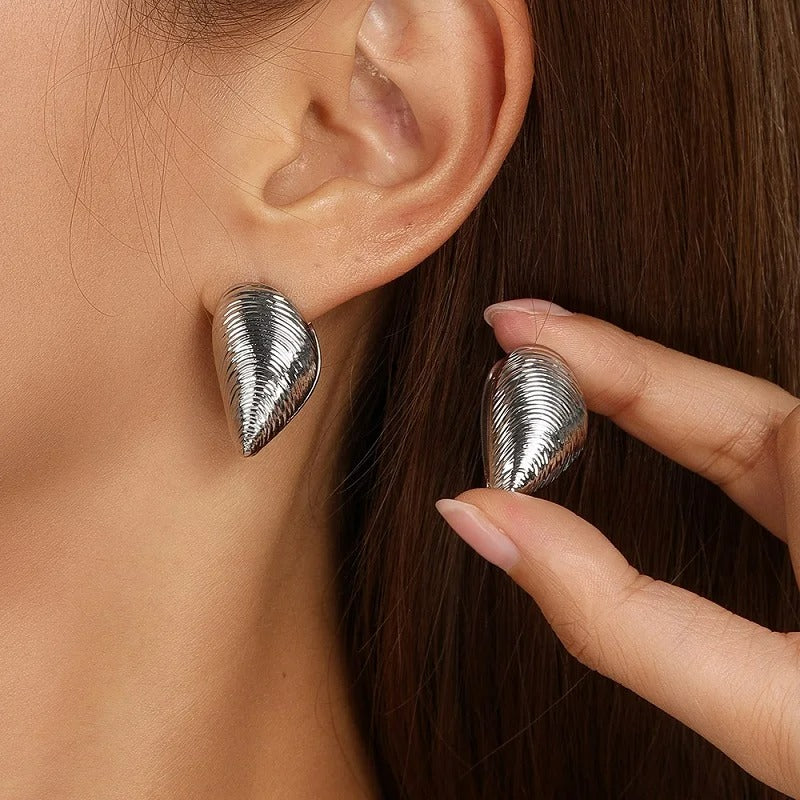 Boucle D'oreille Coquillage Argenté, portée par une femme, elle présente l'autre boucle entre ses doigts à côté de son oreille
