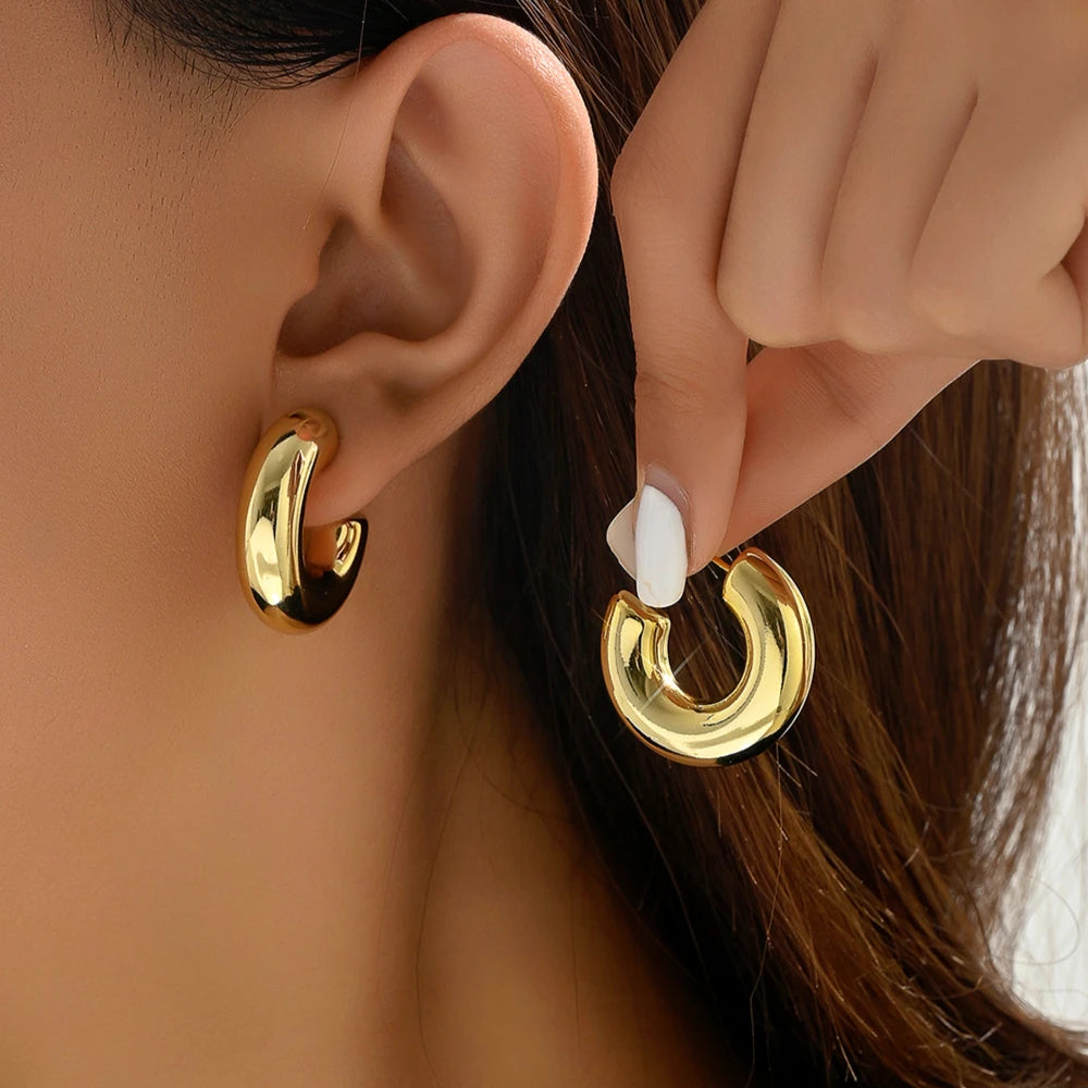 Boucle D'oreille Créole Dorée Épaisse En Acier Inoxydable, portée par une mannequin, l'autre boucle est entre ces doigts, elle la présente à côté de son oreille.