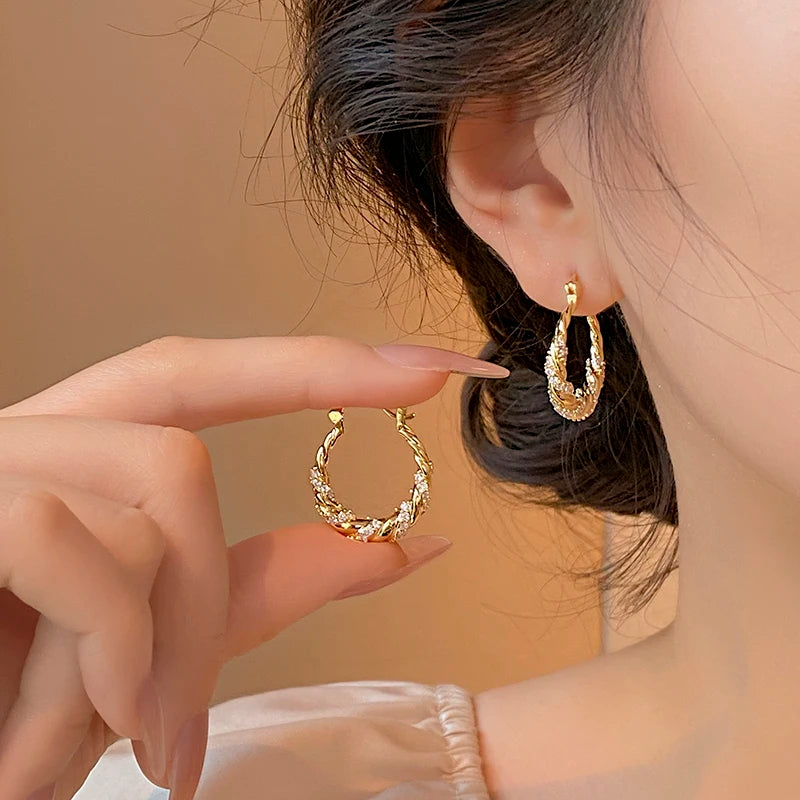Boucle D'oreille Créole Torsadée avec Strass, portée par une femme, elle présente l'autre boucle d'oreille dans ses doigts à côté de celle à son oreille. Image