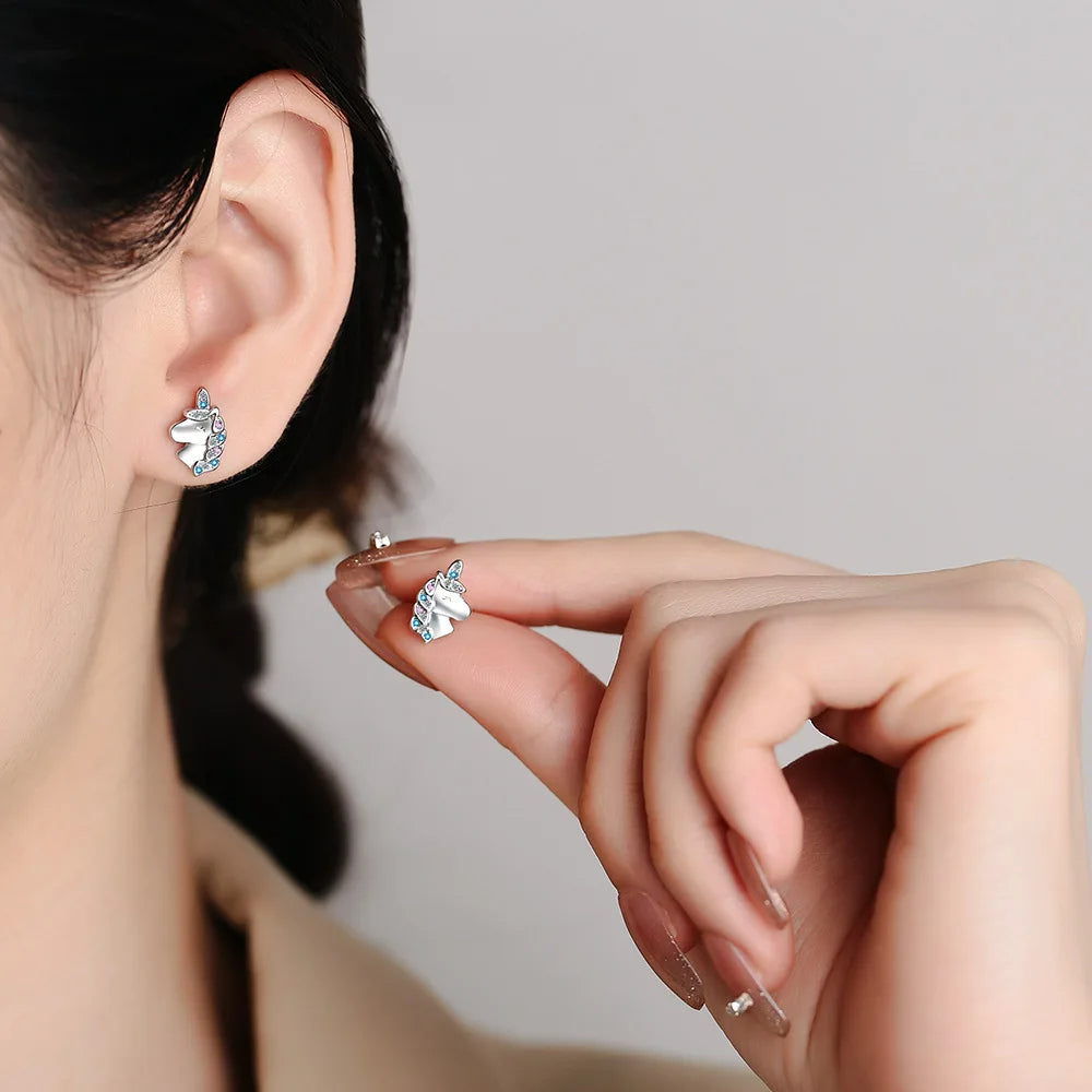 Boucle D'oreille Licorne Strass Roses et Bleus, porté par une femme, elle présente l'autre boucle entre ses doigts à côté de celle à son oreille.