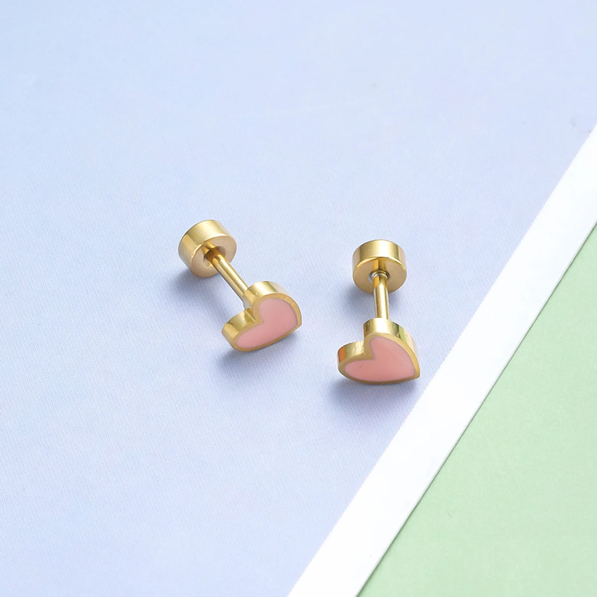 Boucle D'oreille Originale Femme Petit Cœur en Acier Inoxydable dorée et rose, posée sur un papier bleu et vert avec une bande blanche entre les deux couleurs.