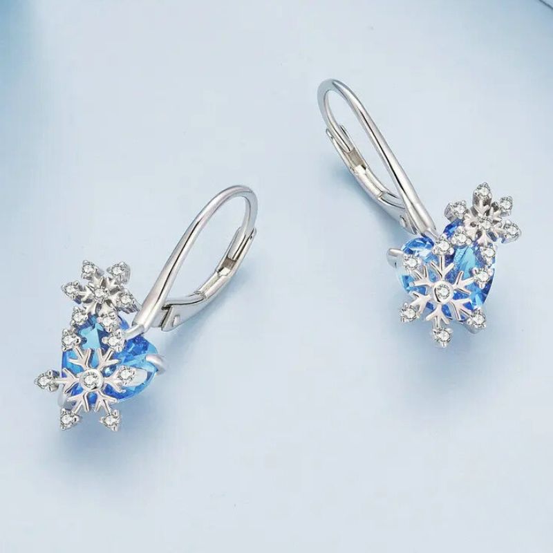 Paire de boucle d'oreilles dormeuse argent flocon et cœur bleu, posée sur un fond bleu pastel.