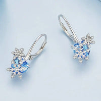 Paire de boucle d'oreilles dormeuse argent flocon et cœur bleu, posée sur un fond bleu pastel.