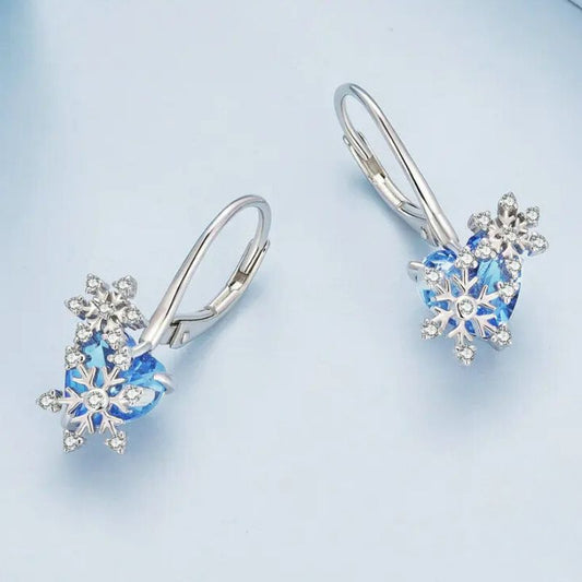 Paire de boucle d'oreilles dormeuse argent flocon et cœur bleu, posée sur un fond bleu pastel.