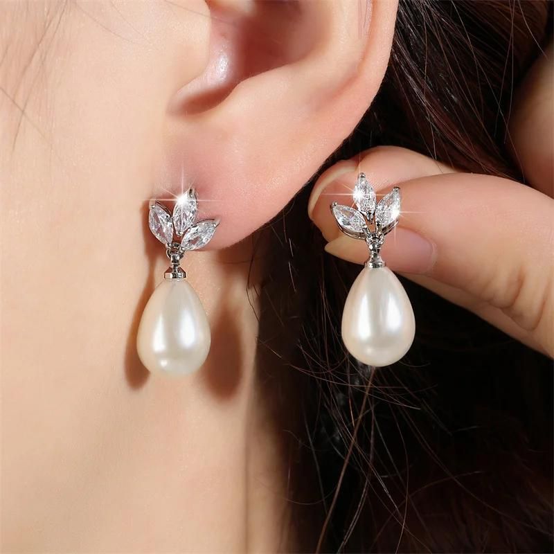 Boucle D'oreille Goutte Perle Nacrée, porté par une mannequin, une boucle accrochée à son oreille, l'autre boucle entre ses doigts qu'elle tient juste à côté. Image