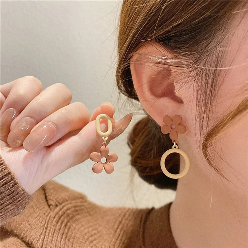 Boucles D'oreilles Asymétriques Fleur Mat, portés par une femme, elle présente l'autre boucle entre ses doigts à côté de celle à son oreille.