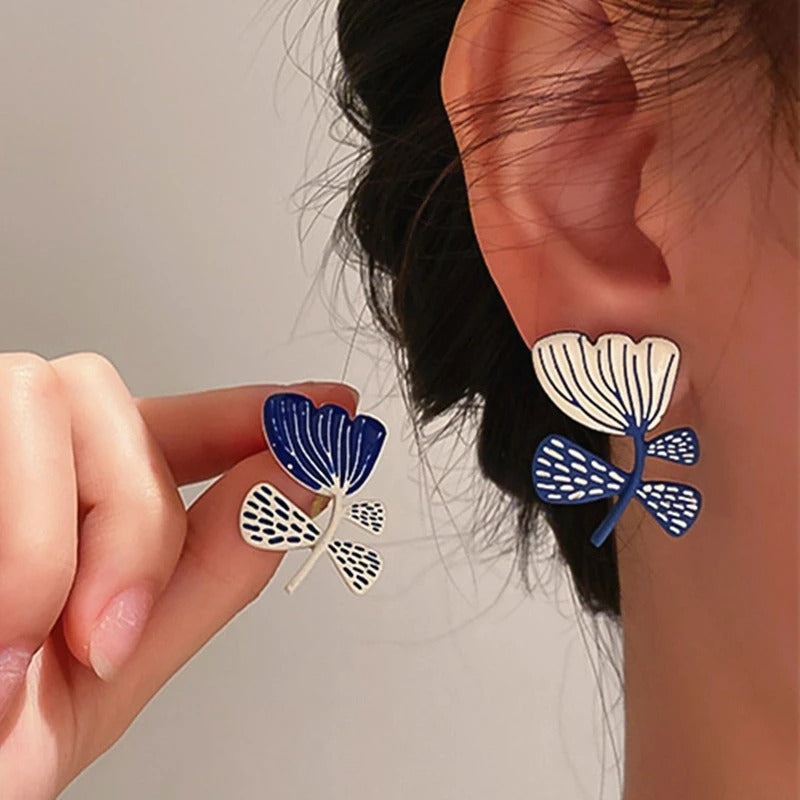 Boucles D'oreilles Asymétriques Fleurs, porté par une femme, une à l'oreille l'autre entre ses doigts présenté à côté de son oreille.