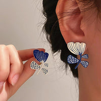 Boucles D'oreilles Asymétriques Fleurs, porté par une femme, une à l'oreille l'autre entre ses doigts présenté à côté de son oreille.